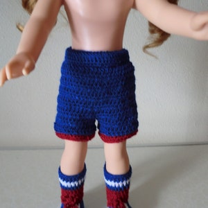 May include: A doll wearing blue and red crocheted shorts and matching socks. The shorts have a red trim and the socks have white stripes.