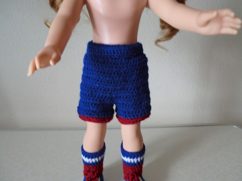 May include: A doll wearing blue and red crocheted shorts and matching socks. The shorts have a red trim and the socks have white stripes.