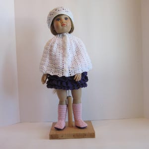 May include: A white crocheted doll cape with a white crocheted hat, a purple ruffled skirt, and pink boots.