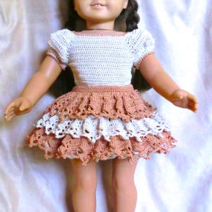 May include: A doll wearing a white and brown crocheted dress with a layered skirt. The doll has brown hair and blue eyes.