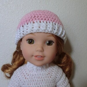 May include: A doll wearing a pink and white crocheted hat. The doll has red hair and brown eyes.