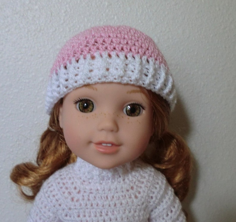 May include: A doll wearing a pink and white crocheted hat. The doll has red hair and brown eyes.
