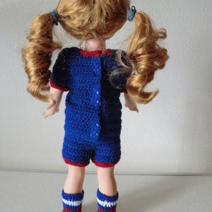 May include: A doll wearing a blue and red crocheted outfit with white accents. The doll has long, curly red hair tied in pigtails.
