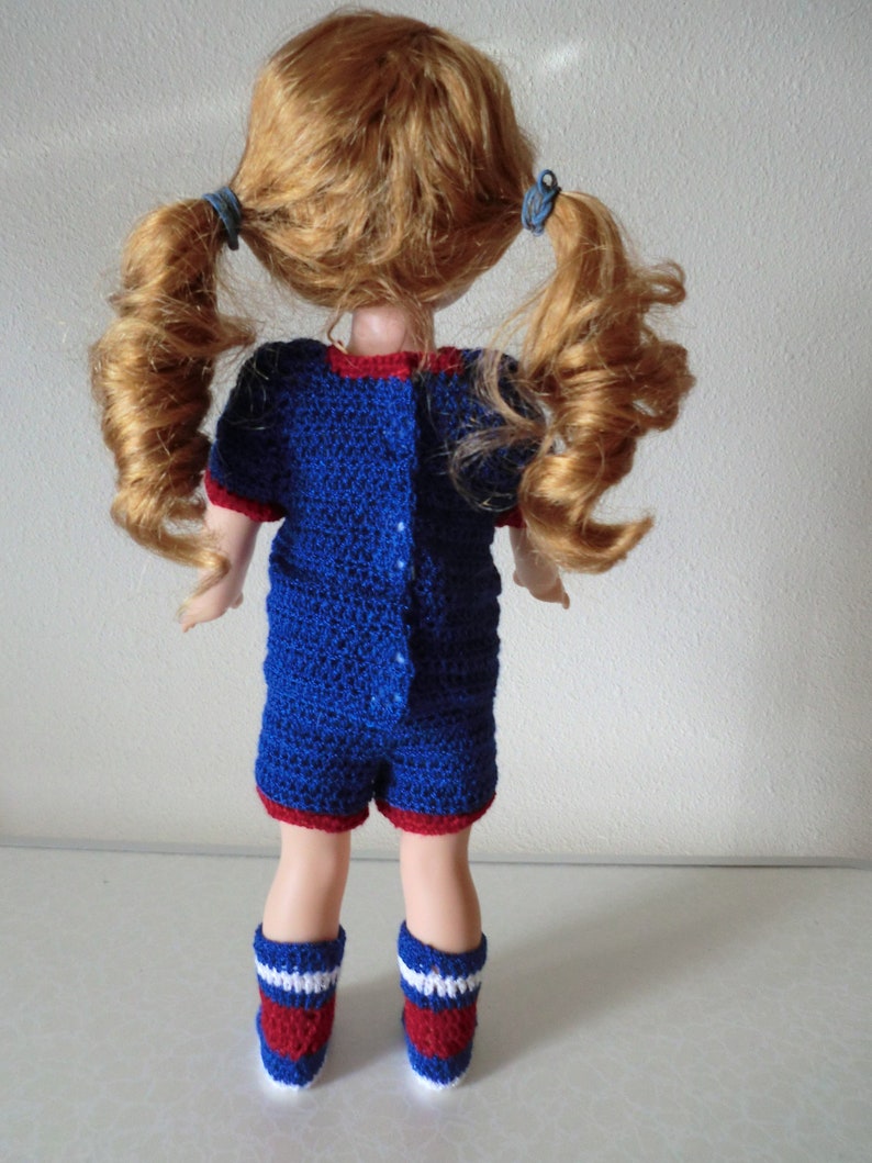 May include: A doll wearing a blue and red crocheted outfit with white accents. The doll has long, curly red hair tied in pigtails.
