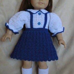 Puede incluir: Una muñeca con un vestido blanco de ganchillo con ribetes azules y una falda azul de ganchillo. La muñeca tiene el pelo castaño y los ojos azules.