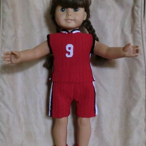 Puede incluir: Una muñeca con un uniforme de baloncesto de crochet rojo y blanco con el número 9 en la parte delantera. La muñeca lleva zapatillas blancas con cordones negros.