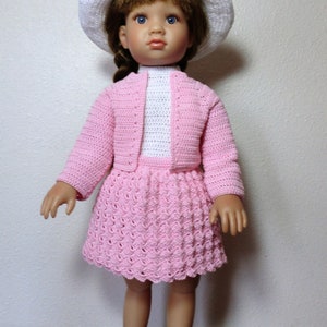 Könnte beinhalten: Ein rosa Häkelkleid und Cardigan-Set mit einem weißen Häkelhut für eine Puppe. Die Puppe hat braunes Haar und blaue Augen.