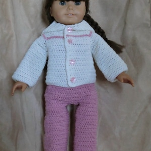 Puede incluir: Una muñeca con un suéter de crochet blanco con ribetes y botones rosas, y pantalones de crochet rosas. La muñeca tiene cabello castaño y ojos marrones.