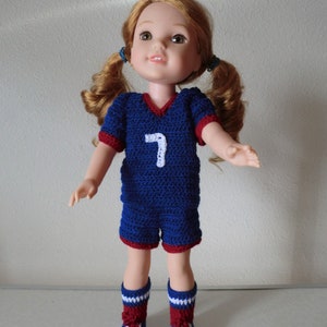 May include: A doll wearing a blue and red crocheted soccer uniform with the number 7 on the front. The doll has long brown hair in pigtails and blue eyes.