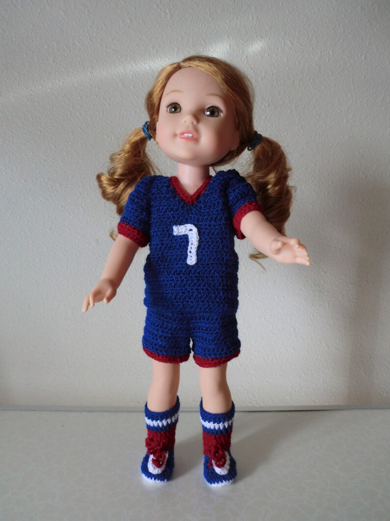 May include: A doll wearing a blue and red crocheted soccer uniform with the number 7 on the front. The doll has long brown hair in pigtails and blue eyes.