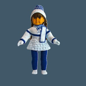 Może przedstawiać: Biały, szydełkowy strój dla lalki z niebieskimi lamówkami. Strój zawiera sweterek, spodnie i czapkę. Lalka ma na sobie białe skarpetki.