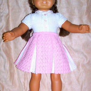 May include: A doll wearing a white crocheted shirt with pink buttons and a pink and white crocheted skirt. The doll has brown hair and blue eyes. The doll is wearing pink and white crocheted socks.