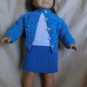 May include: A doll wearing a blue crocheted cardigan, a white crocheted top, and a blue crocheted skirt.