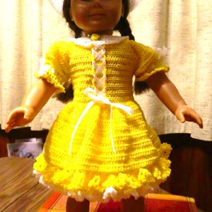 Puede incluir: Un vestido de crochet amarillo con ribete de encaje blanco y un sombrero de crochet blanco. El vestido tiene una cinta blanca atada a la cintura. La muñeca lleva botines de crochet blanco.