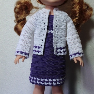 May include: A doll wearing a white crocheted cardigan and a purple crocheted dress with white trim. The doll has long red hair and brown eyes.