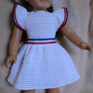 Puede incluir: Un vestido blanco de crochet con ribetes rojo, blanco y azul. El vestido tiene un escote con volantes y una falda completa. La muñeca lleva un sombrero blanco de crochet y zapatos negros con ribetes rojo y blanco.