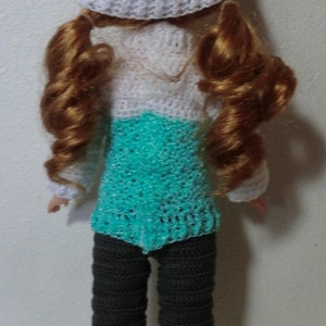 May include: A crocheted doll wearing a teal and white hat, a teal and white sweater, and gray pants with white cuffs.
