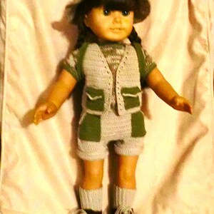 May include: A doll wearing a crocheted outfit with a green vest and shorts. The doll is wearing a brown hat and has brown hair. The doll is standing on a white surface.