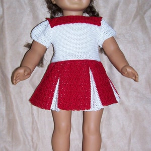 Puede incluir: Una muñeca con un vestido de crochet blanco y rojo con falda plisada. La muñeca lleva zapatos de crochet blanco con detalles rojos.