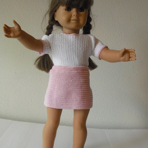 May include: A doll wearing a white and pink crocheted dress with a white crocheted top and a pink crocheted skirt. The doll has brown hair and brown eyes.