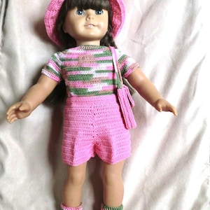 Puede incluir: Una muñeca con un top de crochet a rayas rosa y verde, pantalones cortos de crochet rosa, un sombrero rosa y calcetines de crochet marrones.