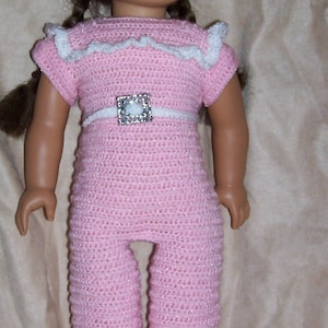 May include: A pink crocheted doll outfit with a white crocheted hat and white shoes. The doll is wearing a white belt with a sparkly buckle.