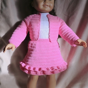 Puede incluir: Un conjunto de muñeca de crochet rosa con un cárdigan, una falda y un dobladillo con volantes. La muñeca lleva zapatos blancos.