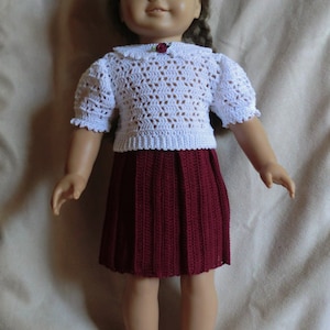 May include: A doll wearing a white crocheted top with a red flower embellishment and a burgundy crocheted skirt.