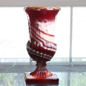 May include: A decorative glass vase with a red and white swirled pattern. The vase has a flared top, a bulbous midsection, and a square base. The colors are a deep red and white, creating a striking contrast. The vase is likely used for displaying flowers or as a decorative piece.