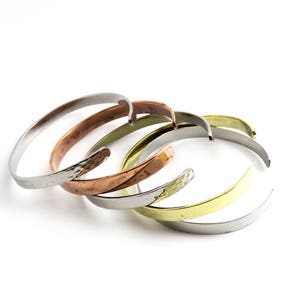 May include: A selection of metal cuff bracelets in silver, copper, brass, and hammered silver finishes. The bracelets are displayed on a white backdrop, highlighting their simple, elegant design. These bracelets are suitable for everyday wear.