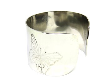 Puede incluir: Un brazalete ancho de color plateado con un diseño de mariposa grabado en la parte delantera. El brazalete tiene un acabado liso y pulido y una forma curva. La mariposa está representada con patrones de alas detallados.