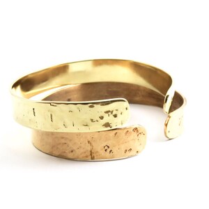 May include: Two gold-coloured metal cuff bracelets. The top bracelet has a hammered texture, while the bottom bracelet has a smoother, lighter finish. The bracelets are open-ended and overlap slightly, showcasing a modern jewellery design.