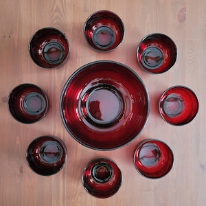 Può includere: Un set di ciotole in vetro rosso rubino e una grande ciotola da portata disposte su una superficie di legno. Le ciotole hanno una finitura lucida e un interno scuro, creando un contrasto sorprendente. Ideale per servire.