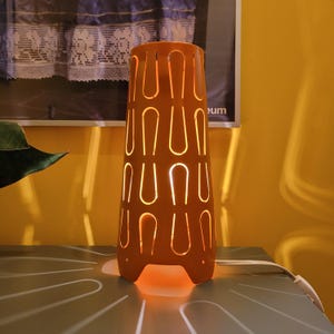 May include: An orange table lamp with a cylindrical shape and a series of vertical, U-shaped cutouts. The lamp is illuminated from within, casting a warm glow. The lamp is plugged into an electrical outlet.
