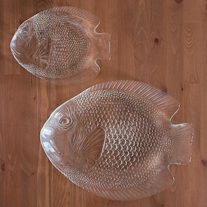 May include: Two clear glass fish-shaped serving platters. The platters have detailed scales, fins, and eye designs. The larger platter is approximately 40 cm long, and the smaller one is about 30 cm long. The platters are on a wooden surface.