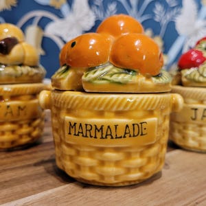 Può includere: Tre vasetti di marmellata in ceramica con coperchi a forma di frutta. I vasetti sono gialli con un motivo a intreccio di cestino e le parole "JAM" e "MARMALADE". I coperchi sono sormontati da frutta arancione e rossa.