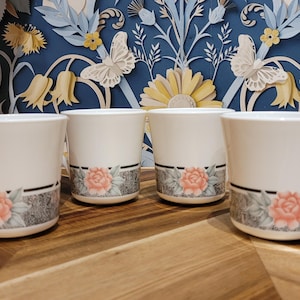 Könnte beinhalten: Vier weiße Keramikbecher mit floralem Design. Jeder Becher hat eine rosa Rose und graues Laub auf einem dekorativen Band. Die Becher haben einen Henkel und stehen auf einer Holzoberfläche.