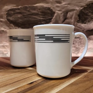 Puede incluir: Dos tazas de cerámica blanca con un diseño de rayas horizontales negras. Las tazas tienen un asa curva y una forma cilíndrica. Las tazas están sobre una superficie de madera.