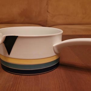 May include: White ceramic saucepan with a long, curved handle. The pan features a black triangle design and horizontal bands of yellow, teal, and navy blue. The saucepan is designed for cooking and serving.