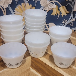 May include: A selection of white glass bowls, some stacked, displayed on a wooden surface. Several bowls have a swan design around the base. The bowls are of different sizes and shapes, ideal for serving or storage.