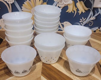May include: A collection of white glass bowls, some stacked, arranged on a wooden surface. Several bowls feature a swan design around the base. The bowls are of varying sizes and shapes, suitable for serving or storage.