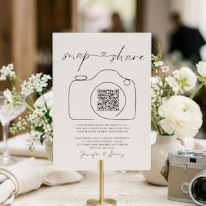 Könnte beinhalten: Ein rechteckiges Schild auf einem goldenen Ständer mit dem Text snap & share, einem Strichzeichnungs-Kamerasymbol mit QR-Code und den Namen Jennifer & Henry. Das Schild enthält Anweisungen zum Hochladen von Fotos in eine Hochzeitsgalerie.