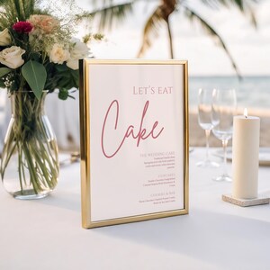 May include: A gold-framed sign with the words "Let's Eat Cake" in a cursive font. The sign is on a white tablecloth, next to a lit candle and a vase of flowers. The background includes the ocean and palm trees.