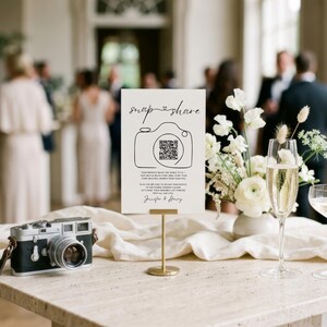 May include: A white rectangular sign on a gold stand featuring the text snap share, a camera illustration with a QR code, and the names Jennifer & Henry. Beside it sits a vintage-style camera, a floral arrangement, and two champagne flutes.