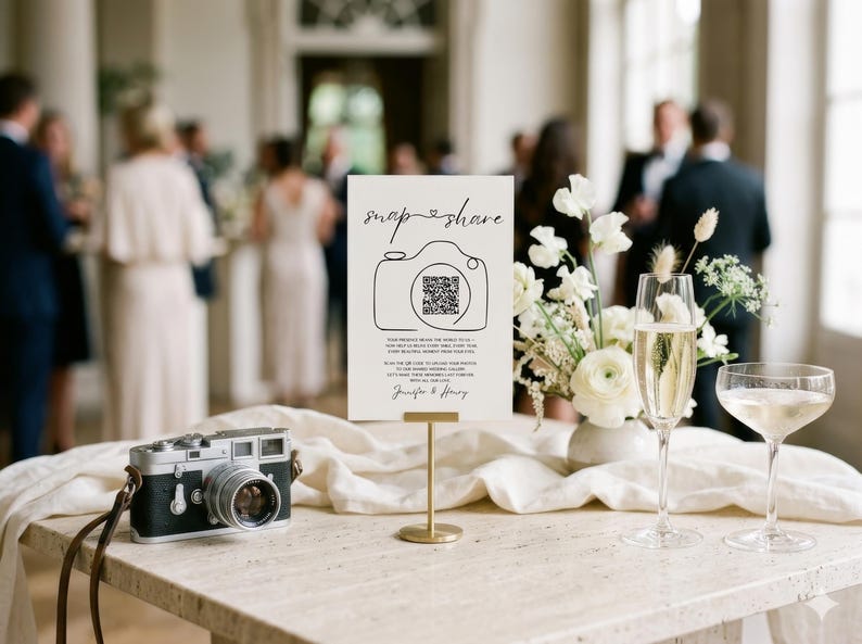 May include: A white rectangular sign on a gold stand featuring the text snap share, a camera illustration with a QR code, and the names Jennifer & Henry. Beside it sits a vintage-style camera, a floral arrangement, and two champagne flutes.
