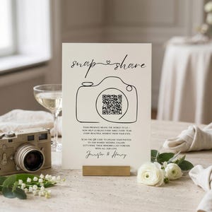 May include: A vertical white wedding sign with black text reading snap share, a line-art camera icon with a QR code in the center, and the names Jennifer & Henry. The sign includes instructions for guests to upload photos to a shared gallery.