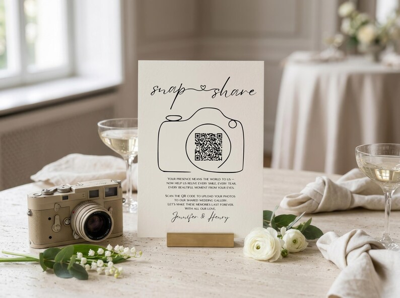 May include: A vertical white wedding sign with black text reading snap share, a line-art camera icon with a QR code in the center, and the names Jennifer & Henry. The sign includes instructions for guests to upload photos to a shared gallery.
