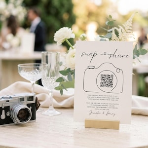 May include: A white rectangular wedding sign with black text and a line-art camera illustration containing a QR code. The sign reads snap share, includes instructions to upload photos to a shared gallery, and is signed by Jennifer and Henry.