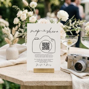 May include: A white rectangular wedding sign with black text and a line-art camera icon containing a QR code. The sign reads "snap & share" and includes instructions to upload photos to a shared gallery, ending with the names "Jennifer & Henry".