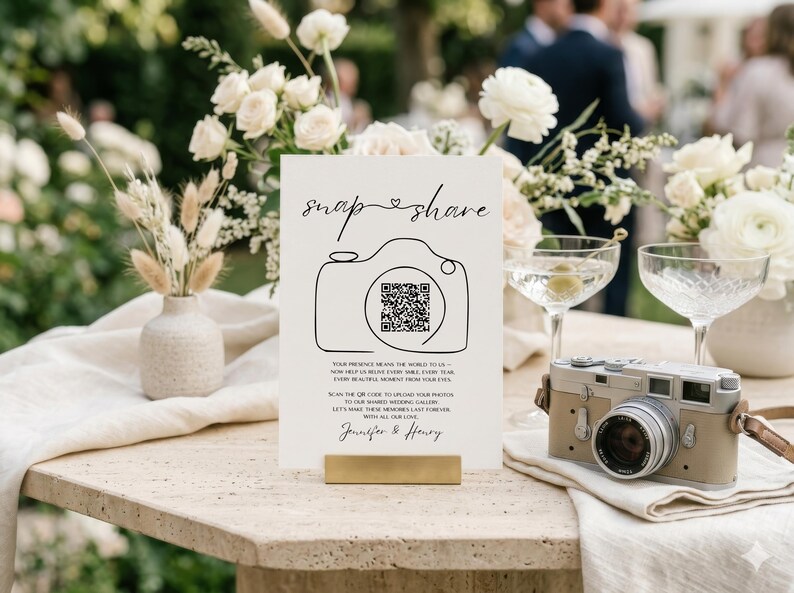 May include: A white rectangular wedding sign with black text and a line-art camera icon containing a QR code. The sign reads "snap & share" and includes instructions to upload photos to a shared gallery, ending with the names "Jennifer & Henry".